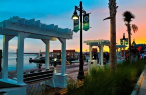 Colorful sunset over the Destin Harbor Boardwalk, with white pergola swings, palm trees, glowing streetlamps, and people walking along the waterfront beside docked boats and calm water.