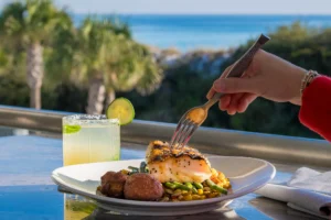 A hand in a red sleeve holds a fork over a plate of grilled fish served on a bed of mixed vegetables with roasted potatoes, set on an outdoor table beside a salted-rim lime cocktail, with palm trees and the ocean blurred in the background.