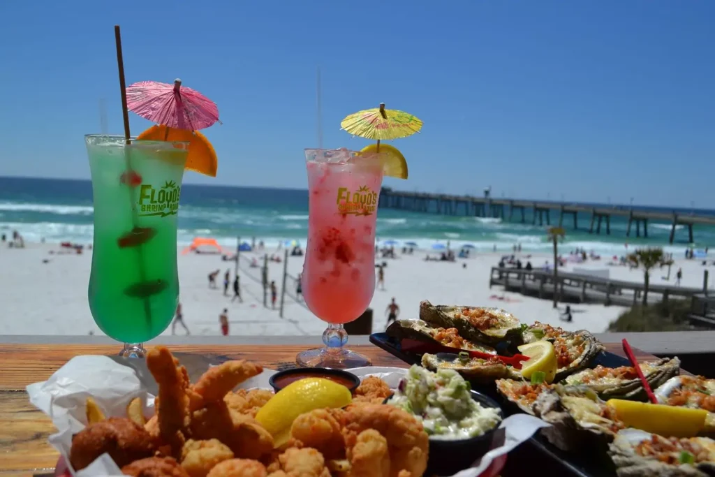 Colorful tropical cocktails and fried seafood platters on a beachfront deck overlooking white sand, emerald-green water, and a long fishing pier.