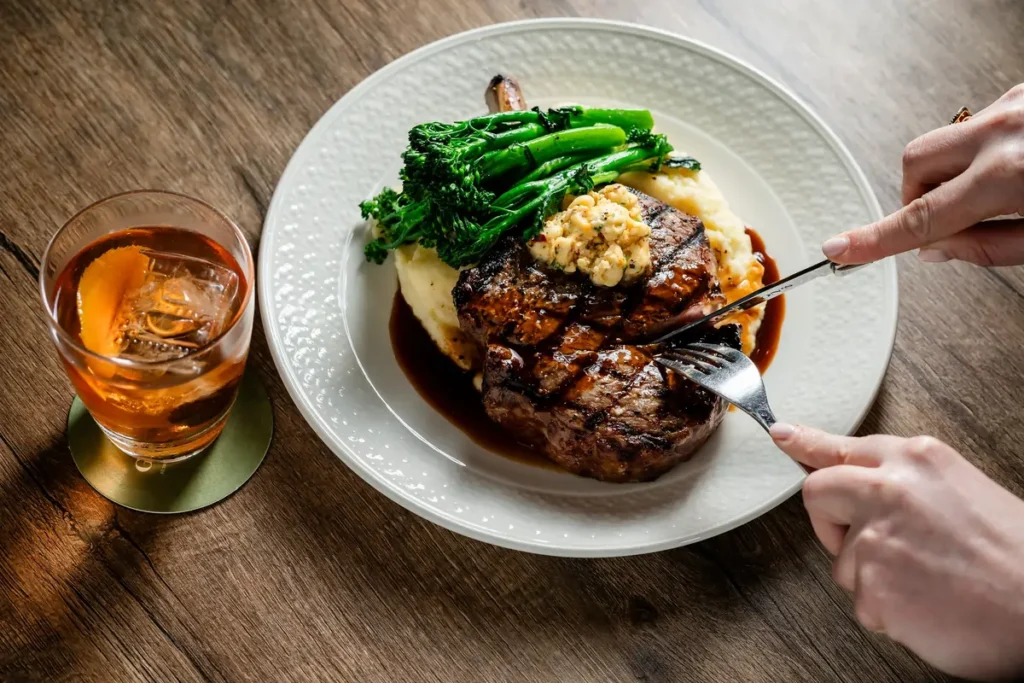 A grilled steak topped with a dollop of seasoned butter is served over creamy mashed potatoes with bright green broccolini on a white plate, while a person cuts into the meat beside a glass of amber-colored cocktail with ice on a wooden table.