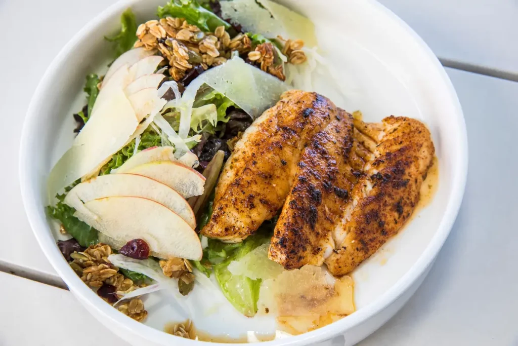 A white bowl filled with a fresh salad of leafy greens, granola clusters, shaved cheese, dried cranberries, and thin apple slices, topped on one side with several pieces of seasoned, grilled fish fillet glistening with pan juices.