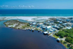 High-angle aerial view of Grayton Beach showing turquoise Gulf waters, a bright white-sand shoreline, a curving coastal dune lake meeting the beach, and a small seaside neighborhood with homes, docks, and pools along the darker inland water.