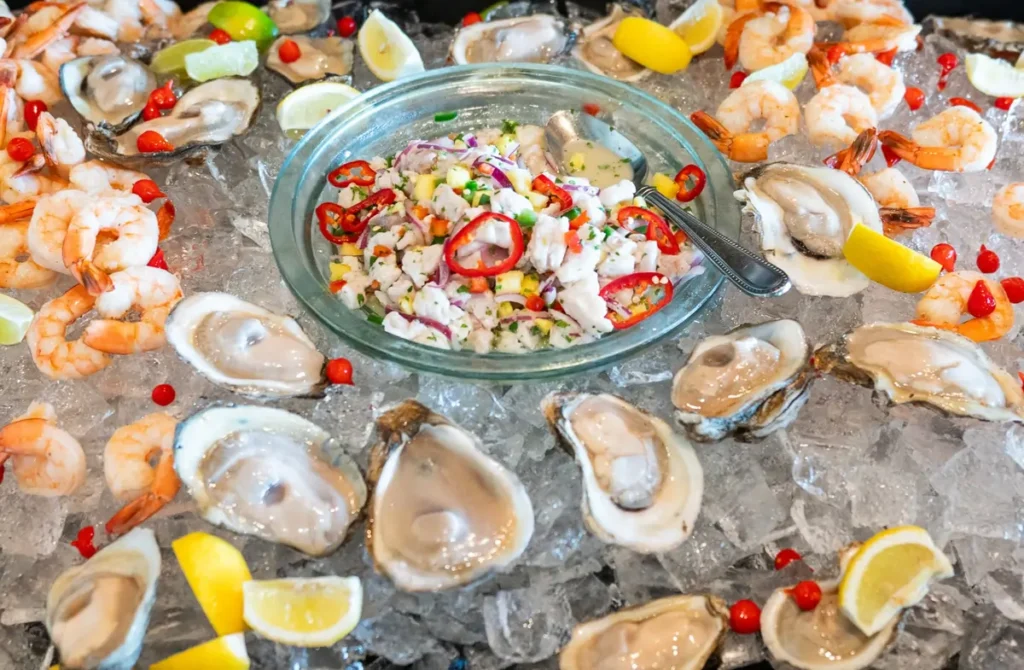 A bed of crushed ice piled with raw oysters on the half shell and chilled shrimp surrounds a glass bowl of colorful ceviche garnished with sliced red chili peppers, lime wedges, and lemon wedges.