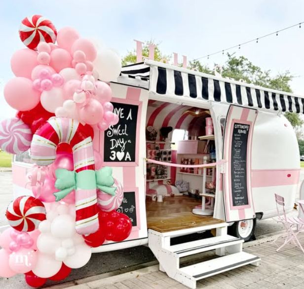 Colorful vintage-style Hello Sugar cotton candy trailer decorated in pink and white stripes, with a huge candy-themed balloon garland, black-and-white awning, chalkboard menus, and an open door revealing jars of sweets inside.