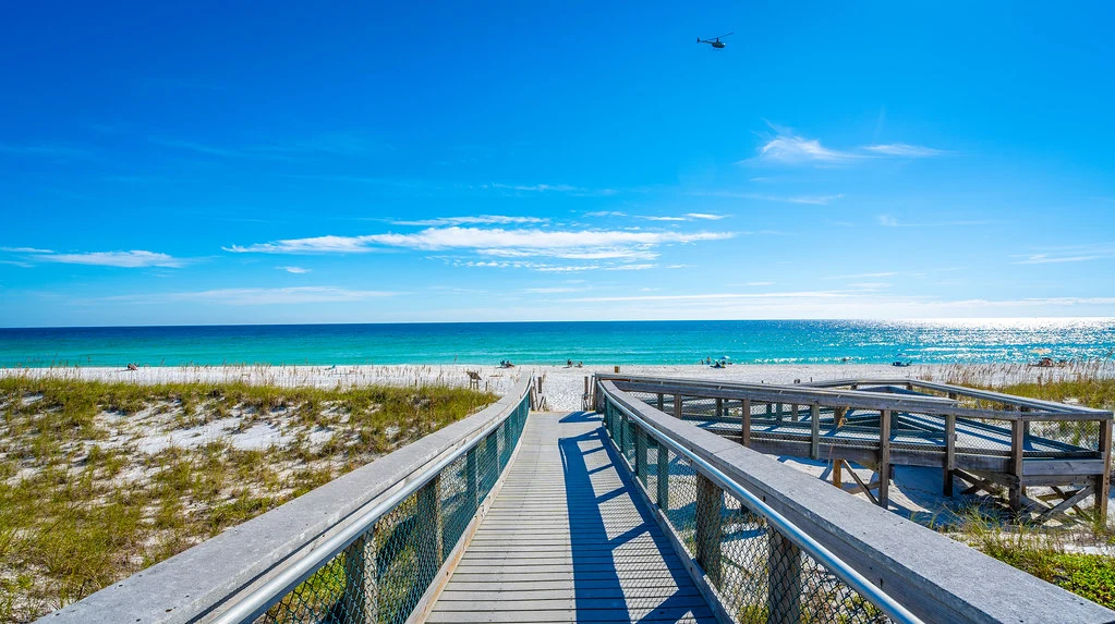 Wooden boardwalk with railings leading over white sand dunes and sea oats to a quiet beach, with turquoise Gulf waters, a bright blue sky, and a small helicopter flying overhead in the distance.