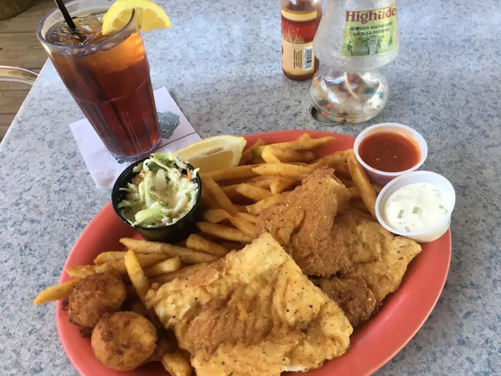 Plate of fried fish, french fries, hush puppies, coleslaw, and dipping sauces with a glass of iced tea on a restaurant table.
