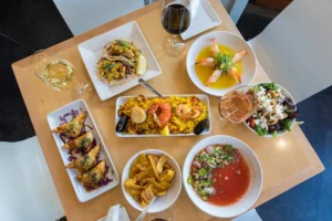 A small wooden table set with a colorful spread of tapas-style dishes, including seafood paella, shrimp in sauce, crispy empanadas, plantain chips, tacos, soup, and a salad, is surrounded by glasses of white, red, and rosé wine.