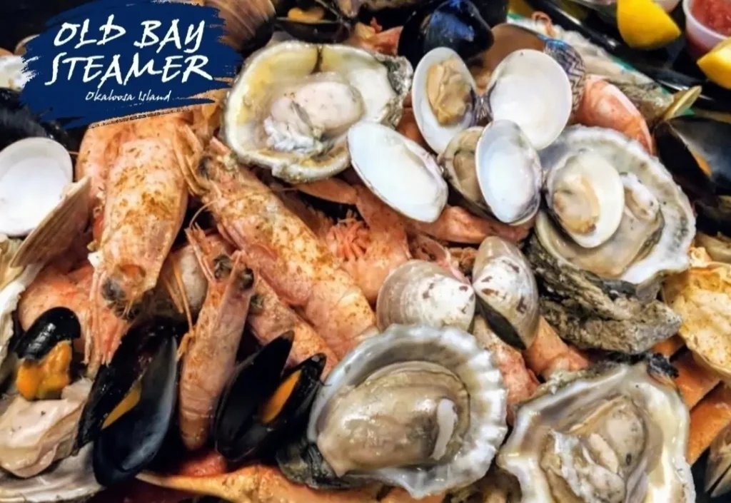 Close-up of a heaping Old Bay–seasoned seafood steamer platter with shrimp, oysters, clams, and mussels, with the Old Bay Steamer logo in the corner.