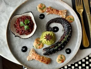 An artfully plated gourmet seafood dish on a large gray plate features a charred octopus tentacle, two seared langoustine tails, a mound of green purée topped with star anise, dollops of pale orange sauce, glossy black sauce circles, and a small bowl of red rice garnished with parsley.