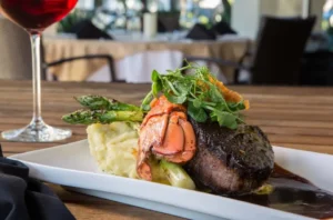 A rectangular white plate holds an elegant surf-and-turf dish: a seared filet mignon with dark crust, topped with greens and paired with a lobster tail, grilled asparagus, and a mound of mashed potatoes, all set over a glossy brown sauce, with a glass of red wine in the blurred background.