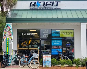 Storefront of Ride It E-Bikes and Paddleboards with large “E-Bikes” window sign, several electric bikes parked outside, a colorful stand-up paddleboard leaning by the entrance, and an “OPEN” sign glowing in the window.