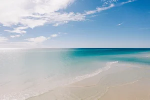 Wide view of Rosemary Beach’s shoreline with glassy, shallow turquoise water lapping onto sugar-white sand under a bright blue sky streaked with wispy clouds.