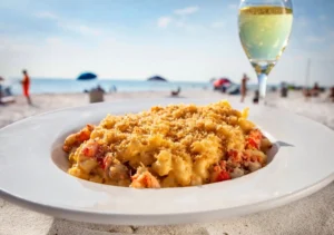 A white, shallow bowl overflowing with creamy macaroni and cheese mixed with chunks of lobster sits on sandy beach ground, topped with a golden breadcrumb crust, while a tall glass of white wine and a blurred shoreline with umbrellas and sunbathers appear in the bright background.