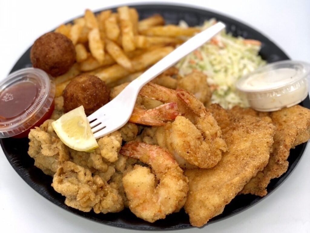 Platter of fried shrimp, fish, and oysters with hush puppies, french fries, coleslaw, and dipping sauces, with a plastic fork resting on top.