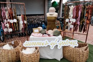 Outdoor pop-up shop featuring racks of colorful women’s swimsuits, woven baskets filled with clothing, a mannequin wearing a fringed top, branded hats on a table, and a large white script sign reading “summer of spivey.”