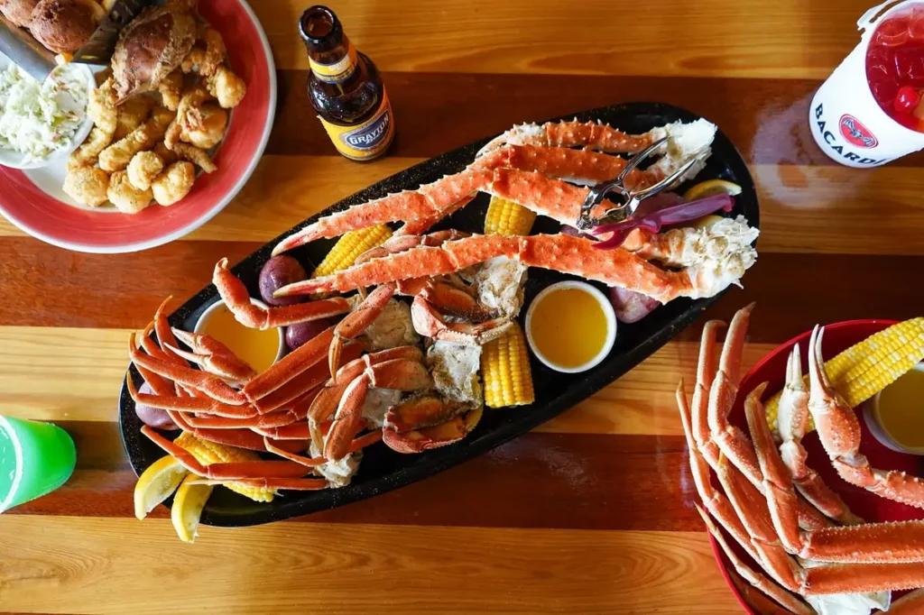 Overhead view of a large crab leg platter with corn, potatoes, and melted butter on a wooden table, surrounded by additional crab and seafood plates and colorful drinks.
