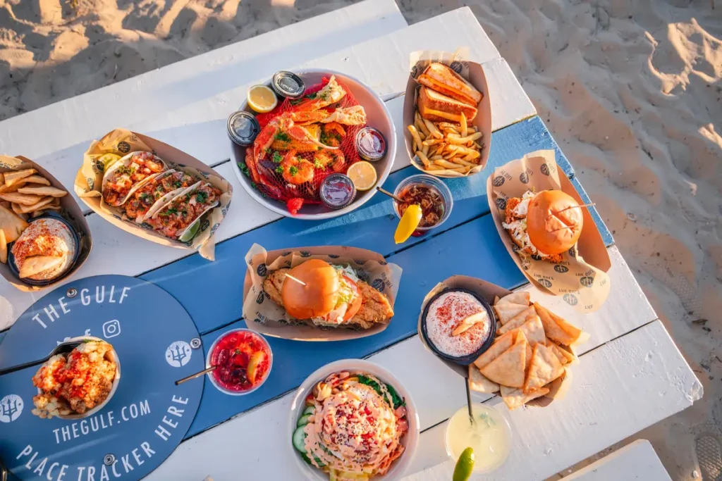 Overhead view of a beachside picnic table covered with seafood tacos, crab legs, burgers, dips, salads, fries, and colorful drinks in the sand at The Gulf.