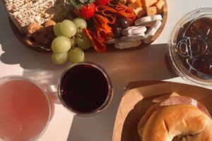 A sunlit spread of snacks and drinks featuring a wooden board with crackers, grapes, strawberries, cheese cubes, and rolled cured meats, alongside a bagel sandwich in a cardboard tray and three different beverages, including red wine and iced coffee in clear cups.