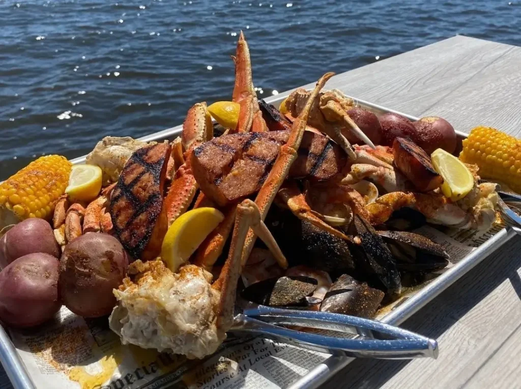 Tray of crab legs, grilled sausage, corn on the cob, potatoes, and shellfish served dockside on a pier overlooking the water.