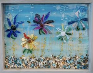 Framed mixed-media artwork featuring bright glass flowers in blue, purple, pink, yellow, and white set against a turquoise background, with textured bubbles in the sky and a border of crushed shells and stones along the bottom.