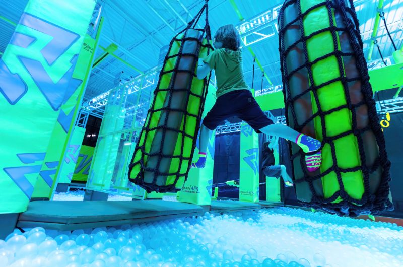 A child in colorful grip socks climbs between two large hanging padded cylinders on a neon-lit indoor obstacle course above a pit filled with white plastic balls at an Urban Air adventure park.
