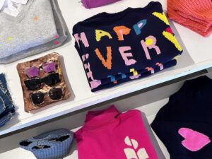 Overhead view of a boutique display table featuring folded sweaters with bright patterns and phrases, including a navy one reading “HAPPY EVERY,” alongside pink and grey knits, sunglasses, jeans, and a woven purse.