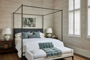 Elegant bedroom with a black four-poster bed dressed in white linens and blue accent pillows, flanked by wooden nightstands with ceramic lamps, a white bench with a blue throw at the foot, textured neutral walls, and tall shuttered windows letting in soft natural light.