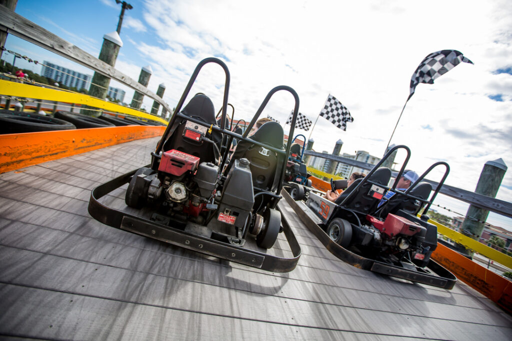 Rear view of several go-karts with roll bars and exposed engines driving up a wooden racetrack ramp, with checkered flags waving and a city skyline visible in the background under a partly cloudy sky.